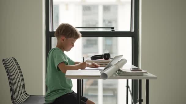 Vit pojke sitter vid bordet med arbetsbok och titta på surfplatta medan uppmärksamt lyssna på lärare online — Stockvideo