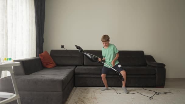 9 ans garçon s'amuser en nettoyant. Un garçon tient un aspirateur comme une guitare et danse avec lui, faisant semblant de jouer tout en écoutant de la musique sur un casque — Video