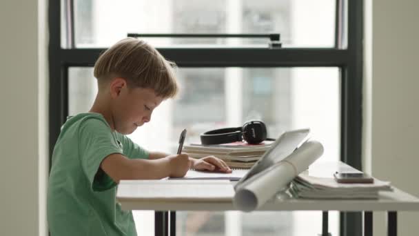 Καυκάσιο αγόρι κάθεται στο τραπέζι με το βιβλίο εργασίας και κοιτάζοντας την οθόνη του tablet, ενώ προσεκτικά ακούγοντας το δάσκαλο σε απευθείας σύνδεση — Αρχείο Βίντεο