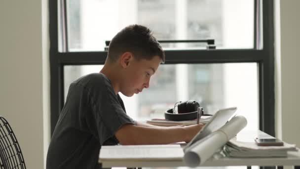 Fehér fiú ül az asztalnál a munkafüzetet, és nézi táblagép képernyőn, miközben figyelmesen hallgat tanár online — Stock videók