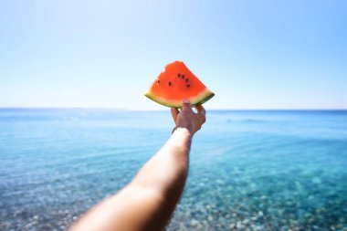 Womans hand holding slice of watermelon on the beach on a sea background. Tropical fruits, healthy food, summer. Sweet, juicy treats in the dish. Healthy eating and lifestyle. Food concept.