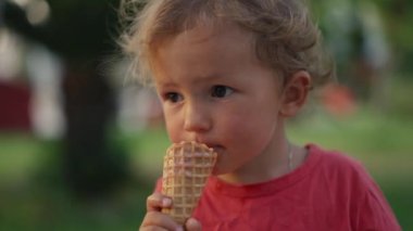 Fair-haired boy eats white cold cream ice cream in waffle cone cup while sitting outside in sunny weather at sunset time. High quality 4k footage