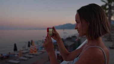 Portrait of woman using modern smartphone for recording video of amazing summer sunset over sea. Focus on mobile screen. Dark atmosphere around. High quality 4k footage