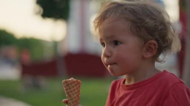 Fair-haired boy eats white cold cream ice cream in waffle cone cup while sitting outside in sunny weather at sunset time. High quality 4k footage