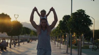 Baba ve oğul gün batımında şehirde yürüyorlar. Ebeveynlerinin omuzlarında iki yaşında bir çocuk oturuyor. Mutlu aile kavramı. Ağır çekimde çekim. Yüksek kalite 4k görüntü