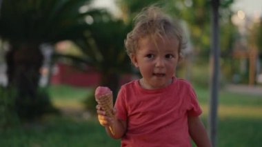 Fair-haired boy eats white cold cream ice cream in waffle cone cup while sitting outside in sunny weather at sunset time. High quality 4k footage