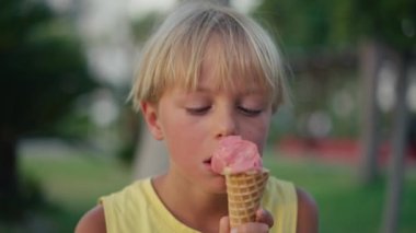 Outdoor portrait of young caucasian boy with blond hair eating ice cream in crispy cone.