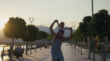 Father and son walking in city at sunset. Two year old kid is sitting on the parents shoulders. Happy family concept. Footage in slow motion. High quality 4k footage