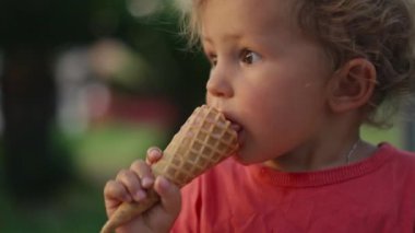 Fair-haired boy eats white cold cream ice cream in waffle cone cup while sitting outside in sunny weather at sunset time. High quality 4k footage