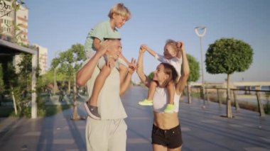 Happy parents and little cute sons on fathers and mothers neck, walking promenade near city lake at sunset. Boy is sitting on fathers shoulders. Happy family spend time together