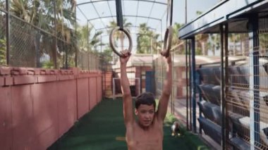 Young athletic boy doing exercise on the gymnastic rings at street workout place. Young athlete doing pull-ups on gymnastic rings. High quality 4k footage