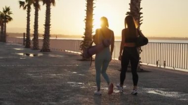 Sport and friendship. Outdoor portrait of two young caucasian women going home after fitness training, carrying fit mats, slow motion. High quality 4k footage
