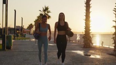 Sport and friendship. Outdoor portrait of two young caucasian women going home after fitness training, carrying fit mats, slow motion. High quality 4k footage