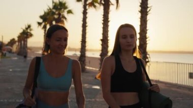 Sport and friendship. Outdoor portrait of two young caucasian women going home after fitness training, carrying fit mats, slow motion. High quality 4k footage