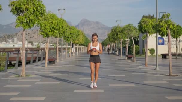アスレチックフィットネススポーツランナーの女の子スポーツ女性が歩くと無線イヤフォンでスマートフォンのリスニング音楽を使用しています 朝の都市公園でワークアウトカーディオ 健康的なライフスタイルを楽しむ若い女性ジョガー — ストック動画