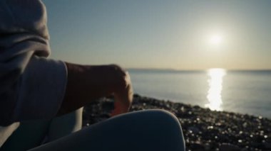 Vücut kıyafetli genç beyaz kadın sakin denizin yanındaki sahilde yoga yaparak dinleniyor. Yakın çekim elleri, Gyan Mudra ve Lotus pozisyonu. Gündoğumu arka planı. Ağır çekim. Yüksek kalite 4k görüntü