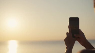 Gün batımında kadın eli cep telefonu tutuyor. Spor elbiseli genç bir kadın deniz aşırı güzel bir gün batımında cep telefonuyla fotoğraf çekiyor. Tatil sırasında akıllı telefondan fotoğraf çekmek.