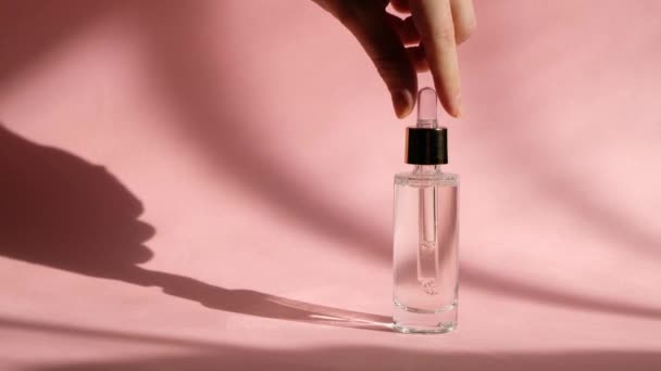 Suero gotero en botella de vidrio transparente sobre fondo rosa y mano de mujer. — Vídeos de Stock