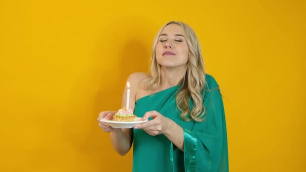 Krásná blond žena slaví a foukání svíčky na dort izolované přes žluté pozadí — Stock video