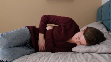 Stresli genç bir kadın yatağa uzanmış, karnını kucaklıyor, karın ağrısı ya da regl sancısı çekiyor. Mide iltihabı, karın ağrısı veya periyod ağrısı.