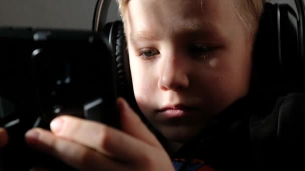 Um rapaz a usar auscultadores com um smartphone nas mãos. Sombras na cara. Criança brinca com telefone, assistindo filme ou mensagens de texto. — Vídeo de Stock