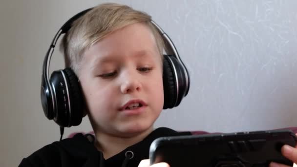Niño usando auriculares con un teléfono inteligente en sus manos. El niño juega con el teléfono, viendo películas o cantando. — Vídeo de stock