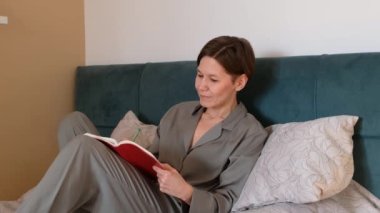 Düşünceli, yaratıcı, genç bir kadın yatakta oturup gelecek planlarını düşünüyor, not defteri günlüğüne notlar alıyor, yaratıcı fikirler yazıyor ve gülümsüyor..