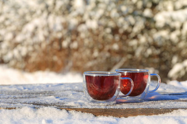 A two glass mugs of tea stand on wooden table on the background of winter landscape.  