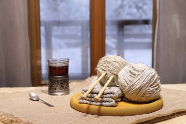 Yünlü iplikler ve örgü örmeler, pencerenin önündeki masanın üzerinde bir bardak çay kış manzarasının arka planında.