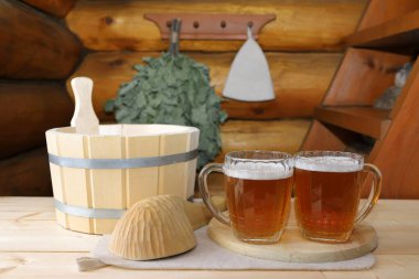 Ahşap bir masada, arka plandaki sauna için aksesuarların yanındaki ahşap bir kapağın üzerinde birkaç bardak bira duruyor üzerinde banyo süpürgesi ve bir şapka asılı kütük bir duvar var..