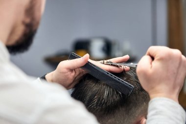 Profesyonel berber, berber dükkanında makas ve tarak kullanarak genç adamın saçını kesiyor. Yakın çekim, seçici odaklanma