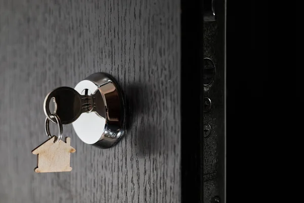 Key with a trinket in the form of a house in the door lock of an open door against the dark background