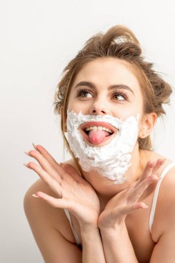 Yüzünde tıraş köpüğü olan güzel, genç, gülümseyen beyaz bir kadının portresi beyaz arka planda dilini dışarı çıkardı.