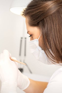 Koruyucu yüz maskesi ve beyaz eldivenli manikür ustası kadın tırnaklarına oje sürüyor.