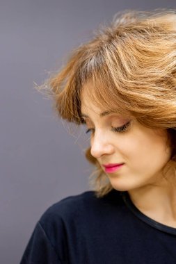 Kızıl kısa kıvırcık saçlı, beyaz, güzel bir kadın koyu arkaplana bakıyor.
