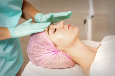 Kozmetik tedavi gören kadın, tıbbi eldivenli kozmetik uzmanı olarak spa salonundaki kadın yüzüne dokunuyor.