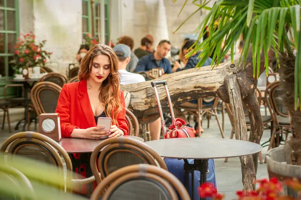 赤のジャケットを着た観光客の若い白人女性は 屋外のカフェでテーブルに座っているスマートフォンを見て — ストック写真