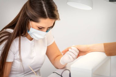 Manikür ustası, beyaz koruyucu maskeli, manikür salonunda ojeli kadın tırnakları boyuyor.