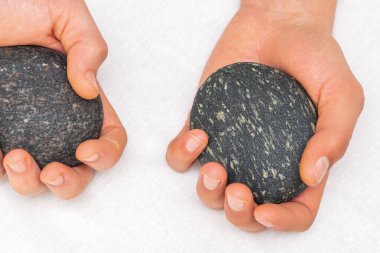 Masörün elleri beyaz arka planda masaj taşları tutuyor.