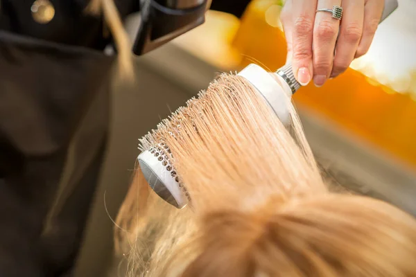 Secagem de cabelo no estúdio de cabeleireiro. estilista de cabeleireira  seca o cabelo com secador de cabelo e escova redonda ruiva de uma mulher em  um salão de beleza.