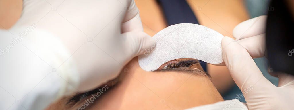The beautician is gluing a patch under her eyes, before the procedure of eyelash extension. Artificial extended eyelashes