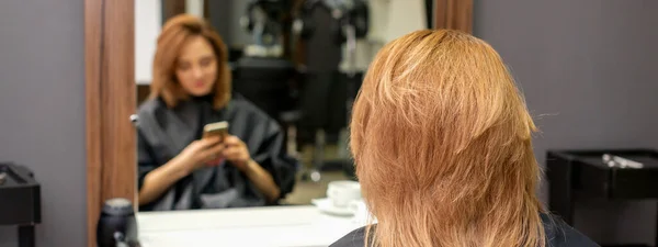 美しい若い赤い髪の女性は彼女のスマートフォンを使用して 鏡の前に座って美容師を訪問するのを待っているテキストメッセージ — ストック写真
