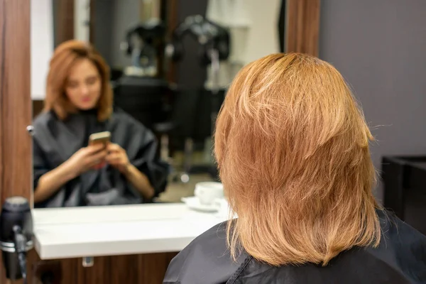 Beautiful young red hair woman using her smartphone and texting sitting in front of a mirror waiting to visit a hairdresser