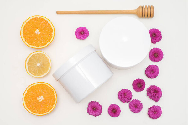 Flat lay composition of white jars with orange slices and flowers on white background, skincare cosmetics, top view