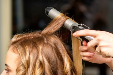 Kuaför, bir güzellik salonunda uzun kahverengi saçlı genç bir kadın için saç maşasıyla bukleler yapıyor.