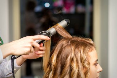 Kuaför, bir güzellik salonunda uzun kahverengi saçlı genç bir kadın için saç maşasıyla bukleler yapıyor.