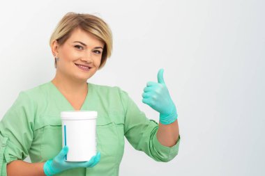 Eldivenli güzellik uzmanı elinde kozmetik ürünü kavanozuyla beyaz arka planda baş parmağını gösteriyor.