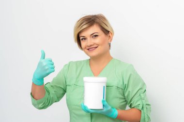 Eldivenli güzellik uzmanı elinde kozmetik ürünü kavanozuyla beyaz arka planda baş parmağını gösteriyor.