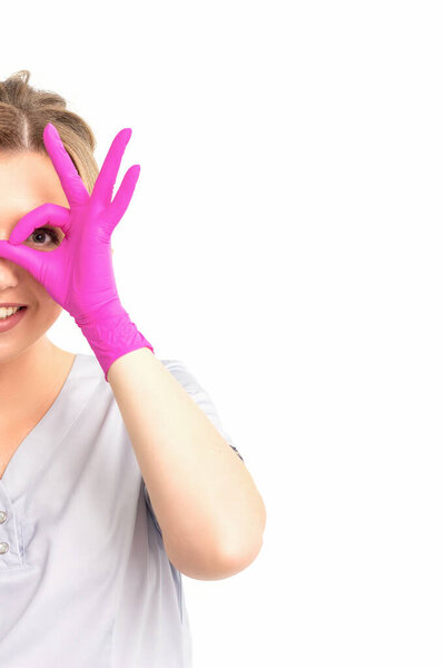 Smiling doctor oculist caucasian woman wearing pink rubber gloves in uniform showing ok sign covering the eye and thumb up gesture against a white background