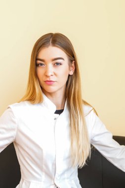 Beyaz önlüklü, sarı saçlı beyaz saçlı beyaz saçlı beyaz saçlı genç bir doktorun portresi. Siyah koltukta oturmuş kameraya bakıyor.